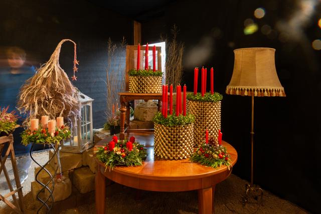 Ein Tisch, auf dem drei Adventskränze mit roten Kerzen stehen. Rechts daneben ist eine braune Stehlampe.