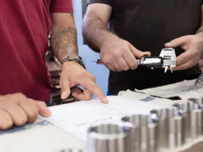 Zwei Mitarbeitende, die sich austauschen. Eine Person zeigt auf einen Plan, die andere miss ein Metallteil ab.